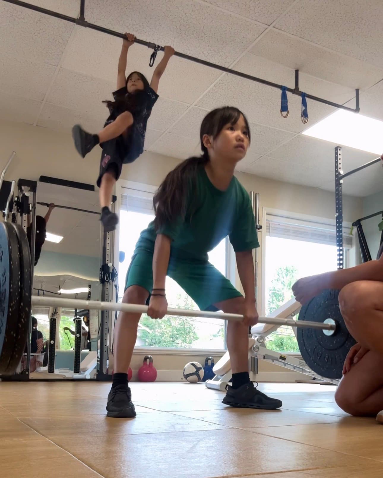 A girl in green deadlifts a barbell while another girl hangs from a pull-up bar.